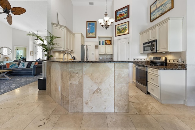 kitchen featuring decorative light fixtures, dark stone counters, a towering ceiling, stainless steel appliances, and ceiling fan with notable chandelier