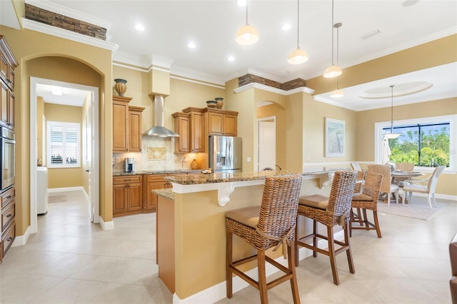 kitchen with light stone counters, stainless steel appliances, wall chimney range hood, pendant lighting, and an island with sink