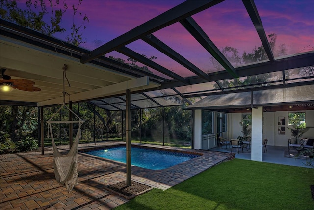 pool at dusk with ceiling fan, a patio area, a yard, and glass enclosure