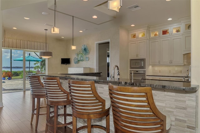 kitchen with built in microwave, stainless steel oven, dark stone countertops, decorative light fixtures, and a kitchen bar