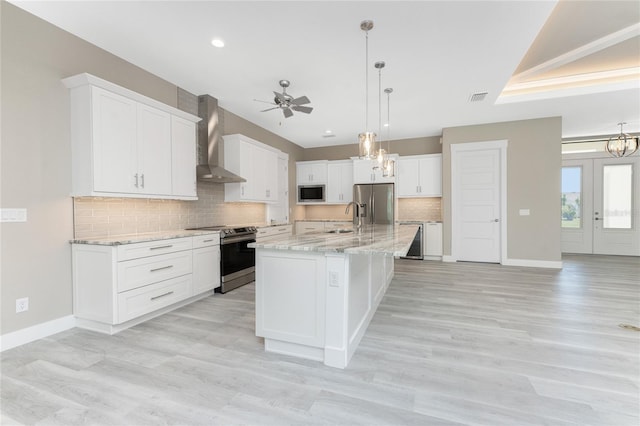 kitchen featuring appliances with stainless steel finishes, ceiling fan with notable chandelier, wall chimney range hood, white cabinetry, and an island with sink