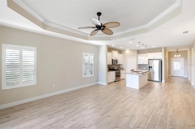 kitchen with ornamental molding, stainless steel appliances, a raised ceiling, hanging light fixtures, and an island with sink