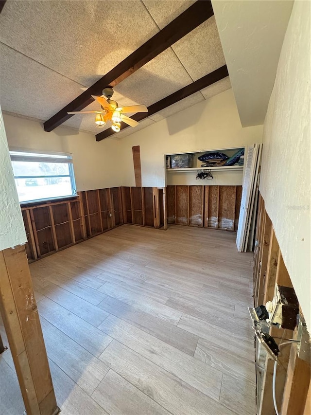 interior space with light wood-type flooring, a textured ceiling, ceiling fan, wooden walls, and vaulted ceiling with beams