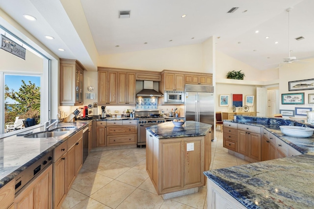 kitchen featuring ceiling fan, wall chimney range hood, built in appliances, dark stone countertops, and a kitchen island