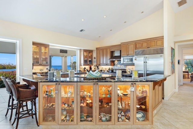 kitchen with built in appliances, wall chimney exhaust hood, a healthy amount of sunlight, and dark stone counters