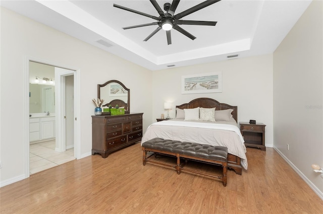 bedroom featuring light wood-type flooring, ensuite bathroom, ceiling fan, and a tray ceiling