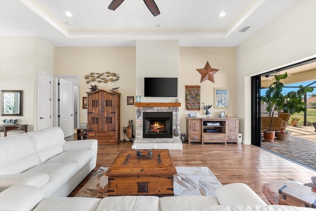 living room featuring a raised ceiling, a stone fireplace, ceiling fan, and light wood-type flooring