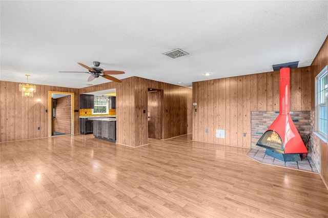 unfurnished living room with ceiling fan with notable chandelier, a textured ceiling, light hardwood / wood-style floors, a wood stove, and wood walls