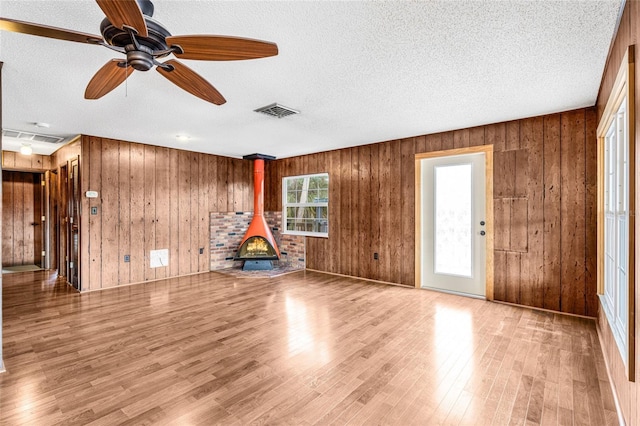 unfurnished living room with a wood stove, ceiling fan, light hardwood / wood-style floors, a textured ceiling, and wooden walls