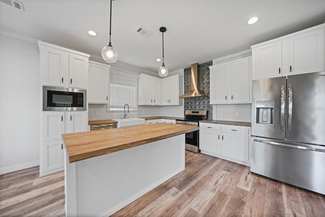 kitchen featuring stainless steel appliances, wall chimney range hood, white cabinetry, hanging light fixtures, and butcher block counters