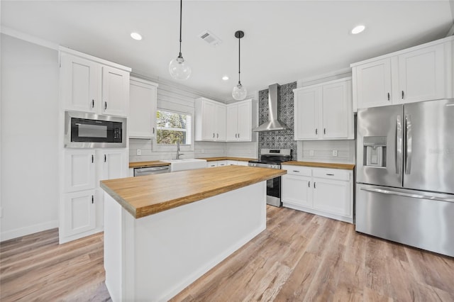 kitchen with white cabinets, butcher block counters, wall chimney range hood, and appliances with stainless steel finishes