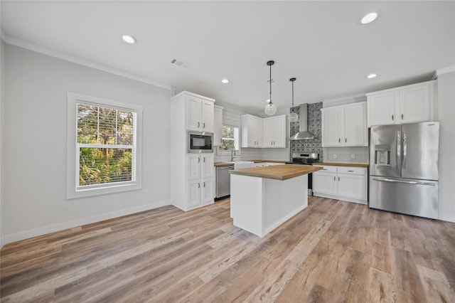 kitchen featuring appliances with stainless steel finishes, wall chimney exhaust hood, pendant lighting, white cabinetry, and butcher block counters