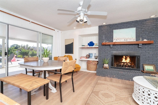 dining area with a ceiling fan, a brick fireplace, light wood-style flooring, and a textured ceiling