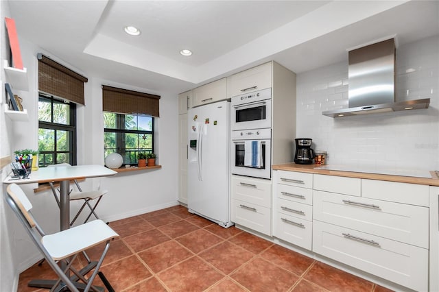 kitchen with white cabinetry, wall chimney exhaust hood, tasteful backsplash, a raised ceiling, and white appliances