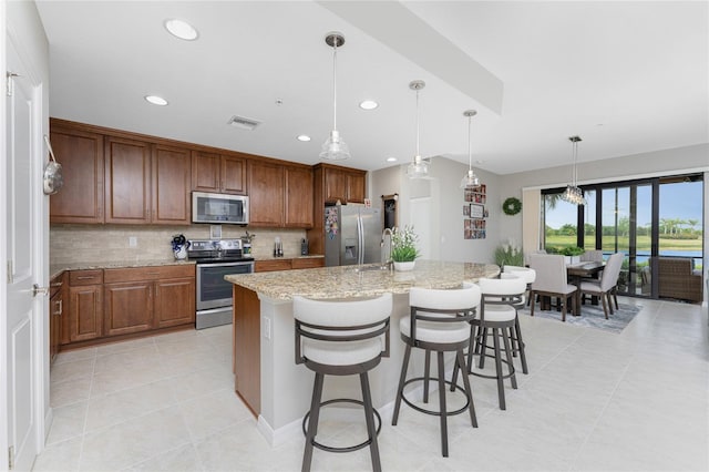 kitchen featuring decorative backsplash, appliances with stainless steel finishes, light stone countertops, pendant lighting, and a center island with sink