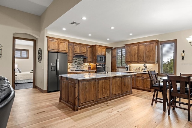 kitchen featuring light stone countertops, a kitchen island with sink, decorative backsplash, black appliances, and light wood-type flooring