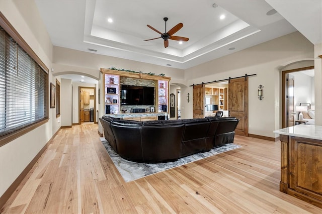 living room with a tray ceiling, a barn door, ceiling fan, and light hardwood / wood-style flooring