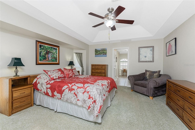 bedroom featuring a raised ceiling, light colored carpet, ensuite bath, and ceiling fan