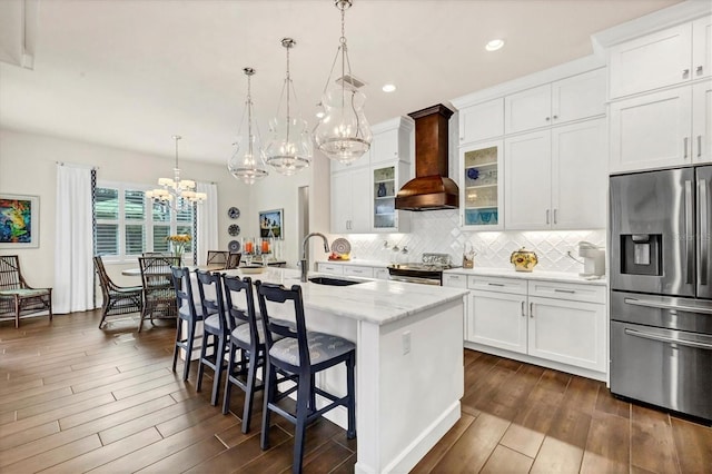 kitchen with custom exhaust hood, stainless steel appliances, sink, hanging light fixtures, and an island with sink