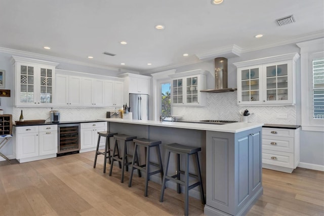 kitchen featuring white cabinets, wall chimney range hood, appliances with stainless steel finishes, a kitchen island, and beverage cooler