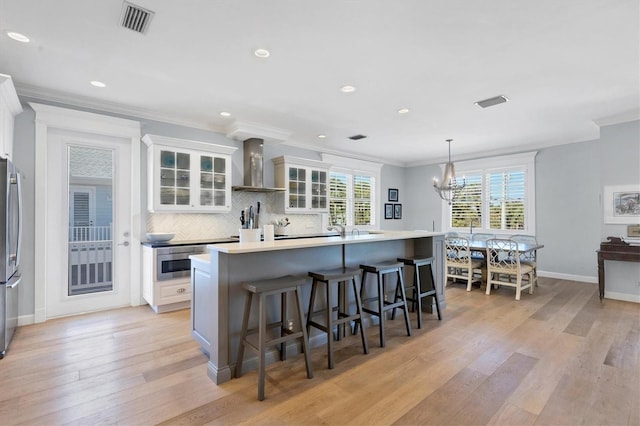 kitchen featuring a center island with sink, white cabinetry, wall chimney range hood, and hanging light fixtures