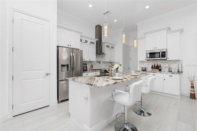 kitchen featuring wall chimney range hood, light stone counters, a center island with sink, white cabinets, and appliances with stainless steel finishes