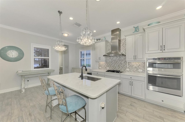 kitchen featuring stainless steel double oven, decorative light fixtures, wall chimney range hood, sink, and a center island with sink