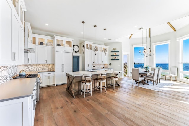 kitchen with backsplash, white cabinets, a water view, hanging light fixtures, and an island with sink