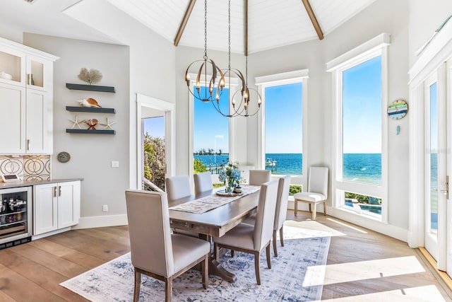 dining space featuring an inviting chandelier, a water view, lofted ceiling with beams, beverage cooler, and light wood-type flooring