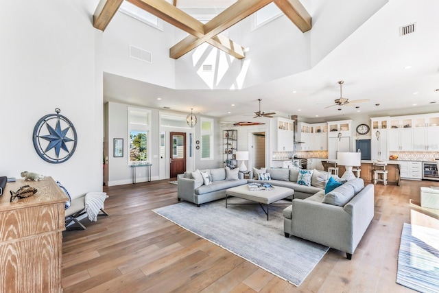 living room with ceiling fan, a towering ceiling, beam ceiling, and light wood-type flooring