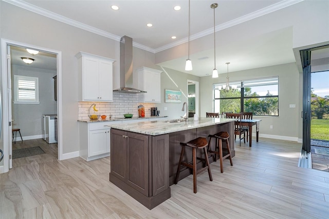 kitchen featuring an island with sink, light stone counters, white cabinetry, wall chimney range hood, and decorative light fixtures