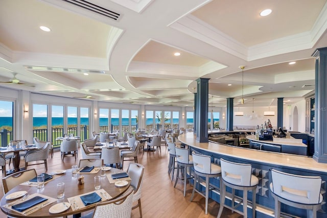 dining area featuring ornate columns, light wood-type flooring, a water view, beam ceiling, and coffered ceiling