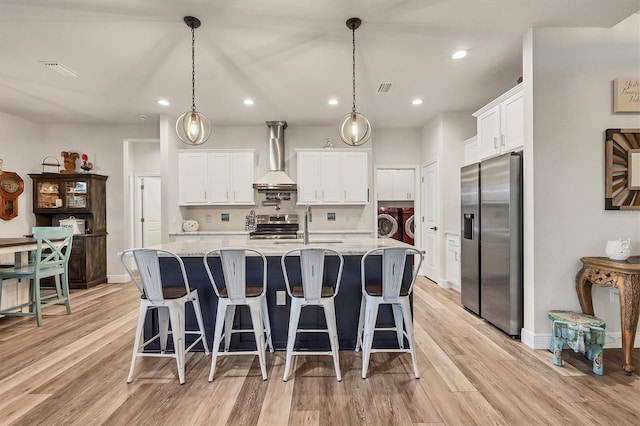 kitchen with appliances with stainless steel finishes, wall chimney exhaust hood, white cabinetry, washing machine and clothes dryer, and a kitchen island with sink