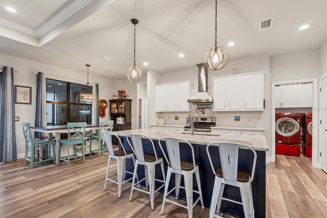 kitchen featuring washer and dryer, light stone countertops, wall chimney range hood, white cabinetry, and a center island with sink