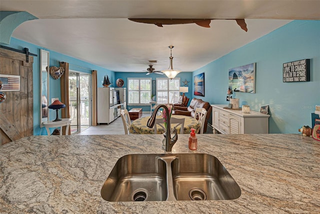 kitchen with sink, ceiling fan, hanging light fixtures, light stone counters, and a barn door