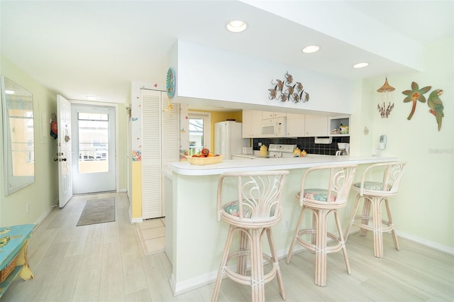 kitchen featuring white cabinetry, backsplash, a kitchen bar, kitchen peninsula, and white appliances