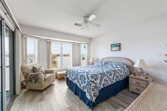 bedroom featuring a textured ceiling, access to exterior, hardwood / wood-style flooring, a water view, and ceiling fan