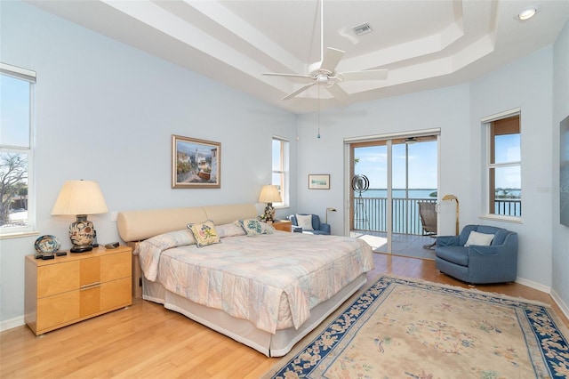 bedroom with access to outside, ceiling fan, a raised ceiling, a water view, and light hardwood / wood-style flooring