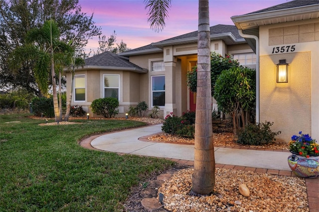 exterior entry at dusk with stucco siding, a shingled roof, and a yard