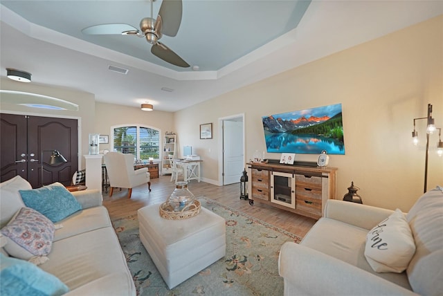 living room with a raised ceiling, ceiling fan, and light wood-type flooring