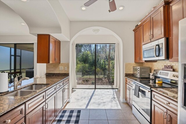 kitchen with light tile patterned floors, stainless steel appliances, a sink, dark stone counters, and brown cabinetry