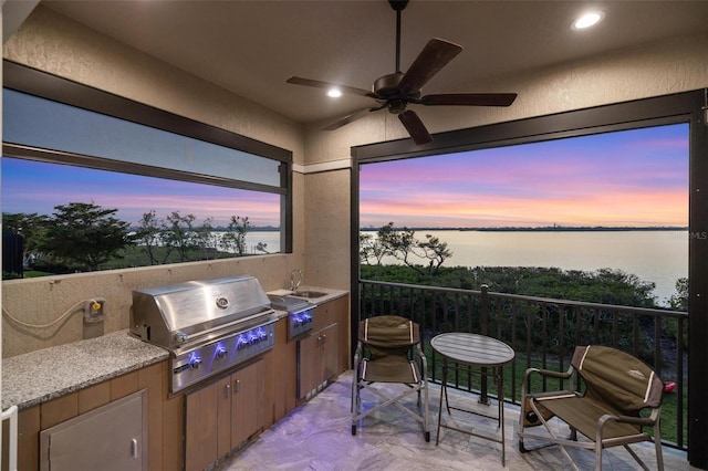 patio terrace at dusk featuring a grill, sink, exterior kitchen, ceiling fan, and a water view