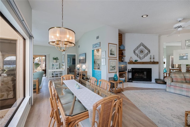 dining room with ceiling fan with notable chandelier, lofted ceiling, a brick fireplace, a textured ceiling, and light hardwood / wood-style flooring