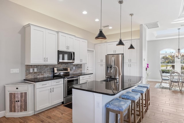 kitchen featuring a kitchen island with sink, white cabinets, appliances with stainless steel finishes, dark countertops, and decorative light fixtures