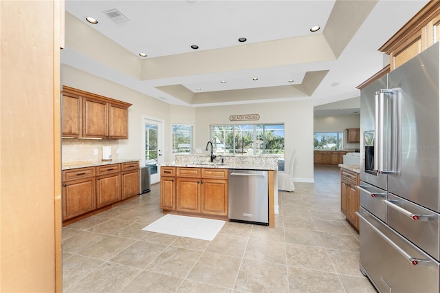 kitchen with appliances with stainless steel finishes, tasteful backsplash, light stone counters, a tray ceiling, and a center island with sink