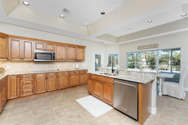 kitchen with sink, appliances with stainless steel finishes, light stone countertops, a center island with sink, and a raised ceiling