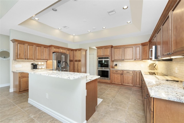 kitchen with a raised ceiling, a center island with sink, light stone counters, and stainless steel appliances