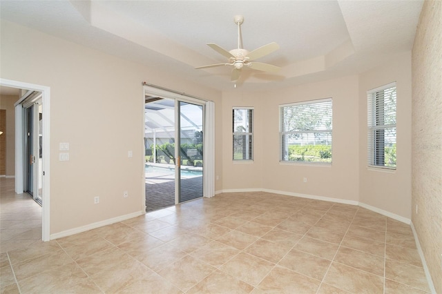 empty room with ceiling fan, a tray ceiling, a healthy amount of sunlight, and light tile patterned flooring