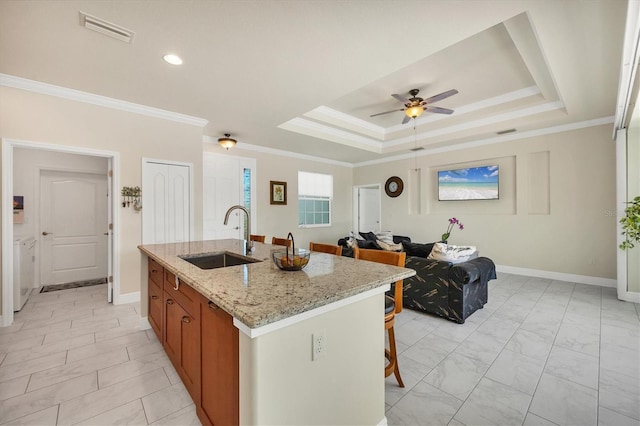 kitchen featuring sink, crown molding, a tray ceiling, light stone countertops, and a kitchen island with sink