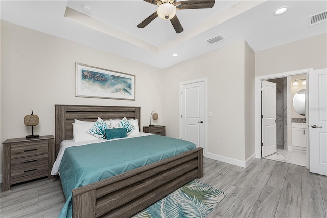 bedroom with ceiling fan, ensuite bathroom, a tray ceiling, and light hardwood / wood-style floors